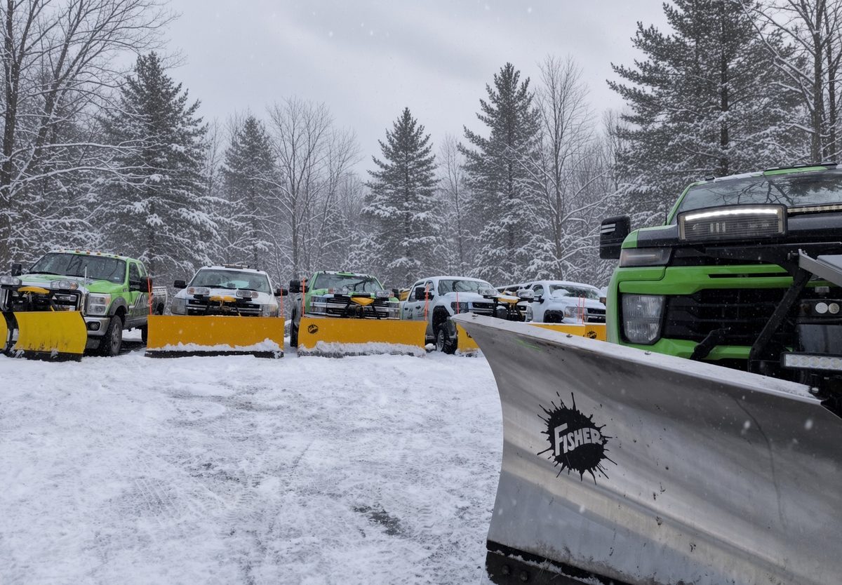 A stainless Fisher plow blade on a green 603 Snow & Ice truck with the rest of the fleet staged behind it in the snow